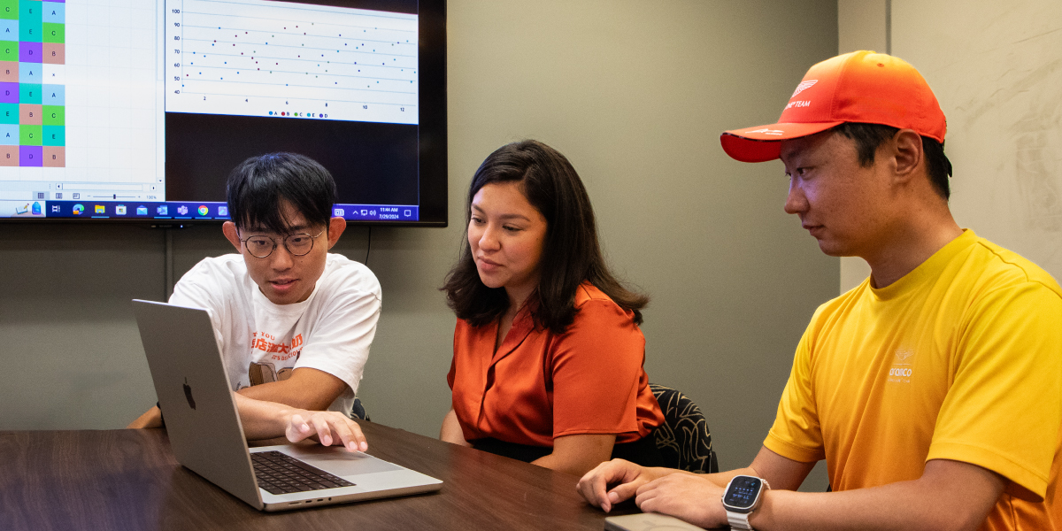 Three people sit at a table around a laptop with a monitor showing statistical analysis on the wall behind them.